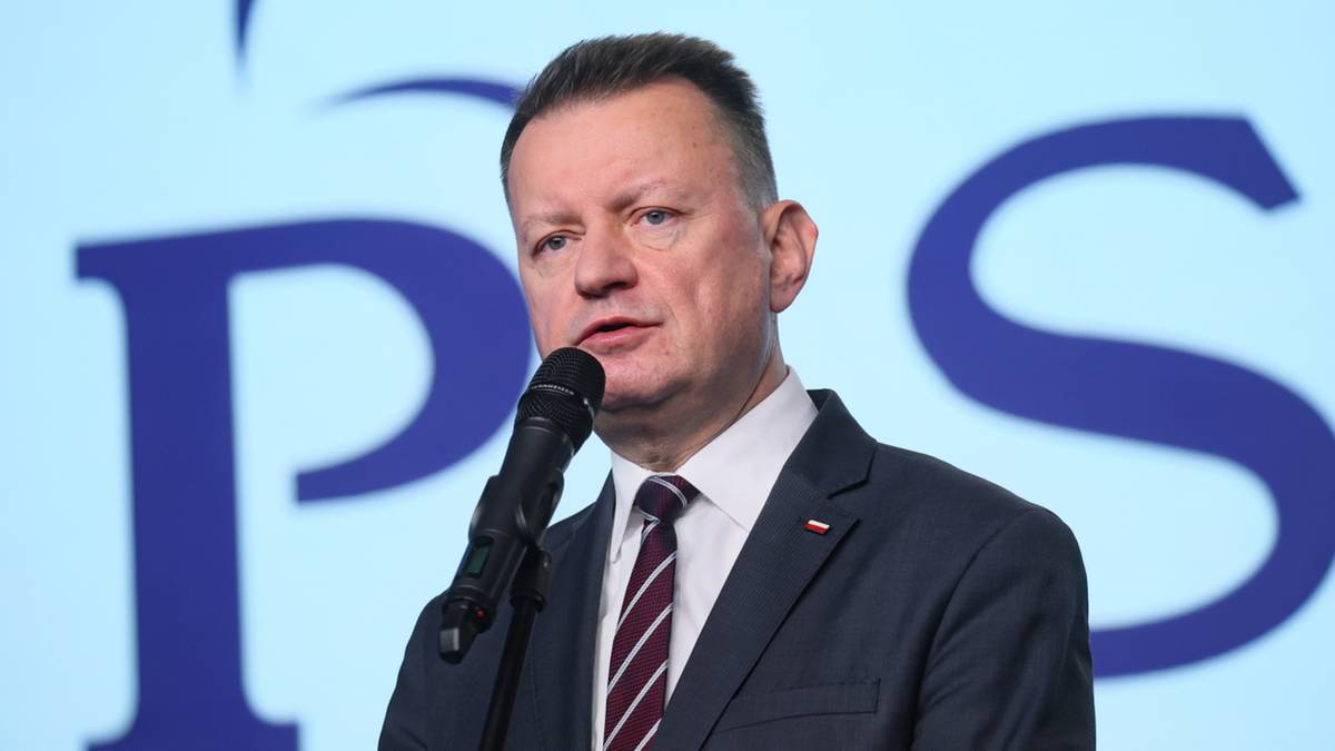 Mężczyzna w ciemnym garniturze i krawacie m&oacute;wi do mikrofonu. W tle widoczne są duże, niebieskie litery tworzące napis "PPS" na jasnym tle.