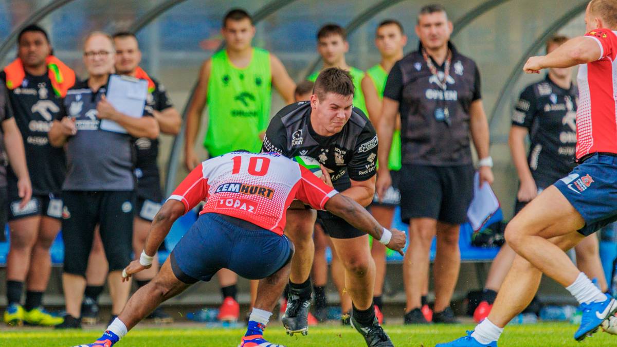 Dwaj zawodnicy rugby w akcji, jeden w białej koszulce z numerem 10 i niebieskimi spodenkami, drugi w czarnej koszulce. W tle widać innych zawodników i kibiców.