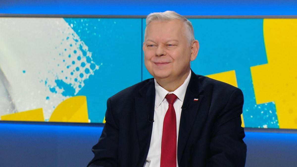 Mężczyzna w garniturze i czerwonym krawacie siedzi przed kolorowym tłem.