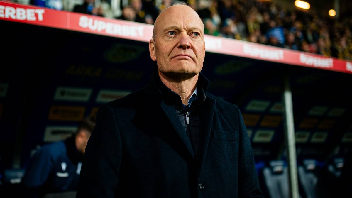 Trener Lecha Poznań Frederiksen w ciemnym płaszczu, patrzący przed siebie z poważną miną, na tle stadionu.