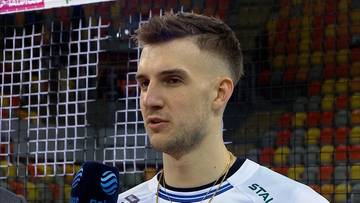 Młody mężczyzna o jasnej karnacji, krótkich, ciemnych włosach i zaroście, ubrany w białą koszulkę sportową z niebieskimi elementami. Patrzy w bok, lekko zmrużonymi oczami. W tle widoczna siatka do siatkówki i trybuny wypełnione pomarańczowymi siedzeniami.