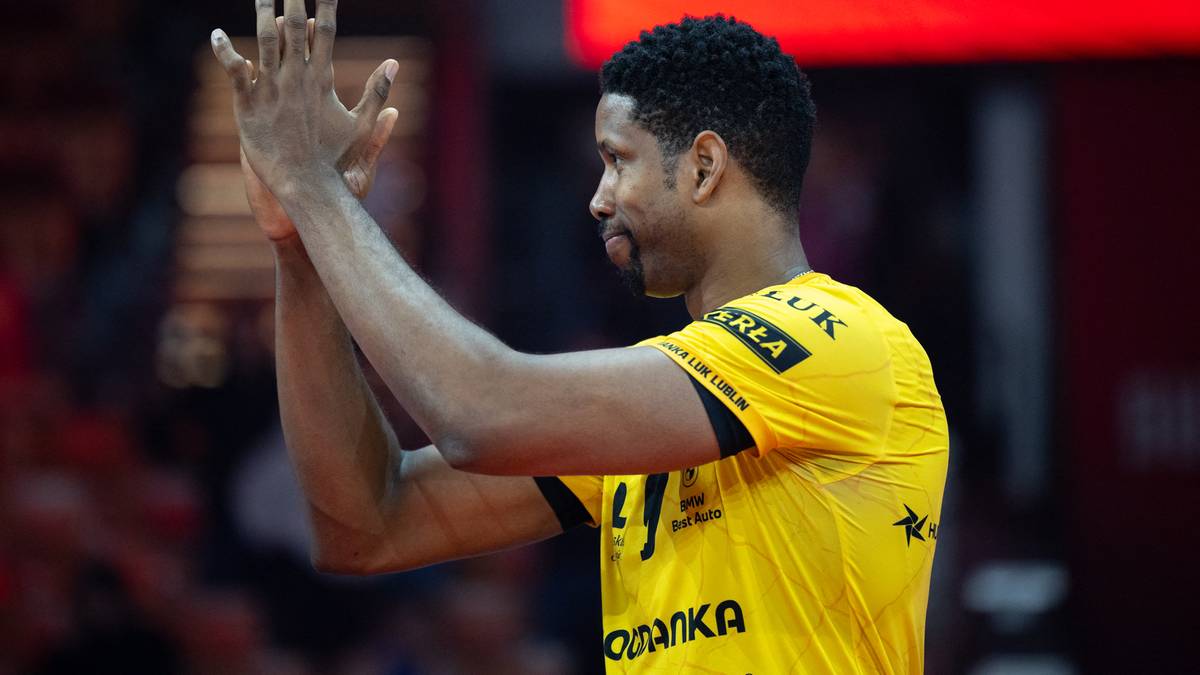 Siatkarz w ż&oacute;łtej koszulce z kr&oacute;tkim rękawem, z czarnymi wstawkami, zaciska dłonie na wysokości klatki piersiowej, patrząc w bok.