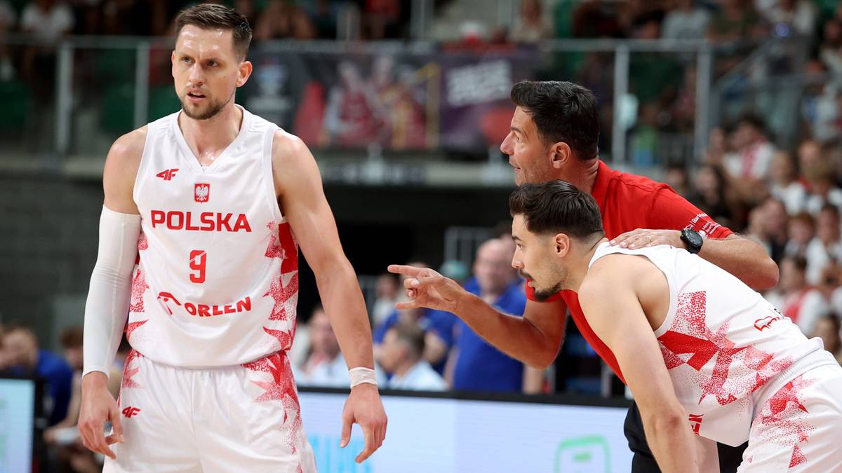 Trzech mężczyzn w strojach koszykarskich. Po lewej zawodnik z numerem 9 na koszulce z napisem "POLSKA" i herbem Polski. Pośrodku mężczyzna w czerwonej koszulce, wskazujący palcem w prawo. Po prawej drugi zawodnik w białej koszulce z czerwonymi elementami.