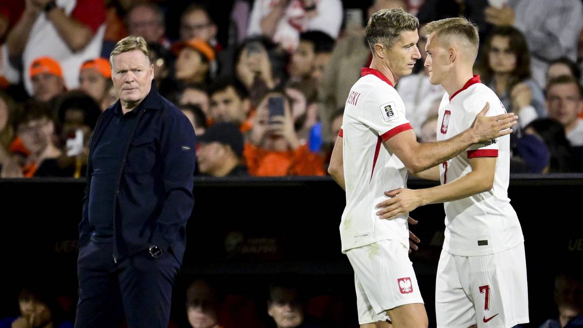 Trzech mężczyzn na boisku piłkarskim. Po lewej starszy mężczyzna w ciemnej kurtce. Po prawej dwóch młodych mężczyzn w białych strojach piłkarskich.