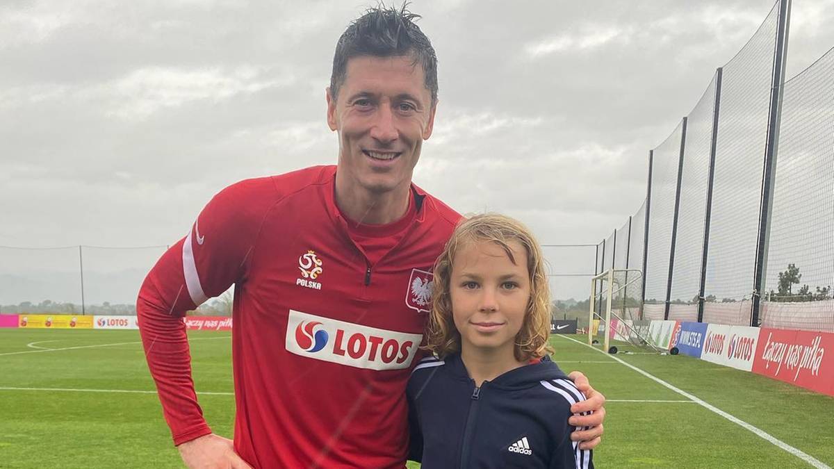 Robert Lewandowski w czerwonej koszulce reprezentacji Polski pozuje do zdjęcia z młodym chłopcem na tle boiska piłkarskiego.