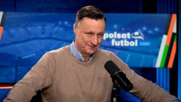 Mężczyzna w brązowym swetrze i koszuli w niebieskie paski, stojący przed mikrofonem, z uśmiechem na twarzy.