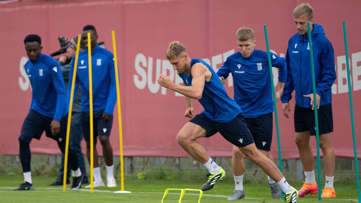 Piłkarze Lecha Poznań w niebieskich strojach treningowych biegają między tyczkami, wykonując ćwiczenia zwinnościowe. W tle czerwona ściana z logo klubu i napisami.