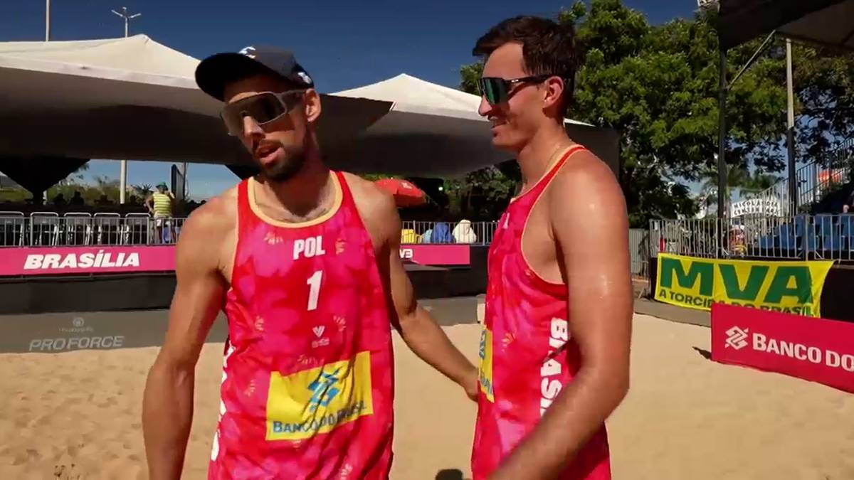 Dw&oacute;ch mężczyzn w czerwonych strojach sportowych do siatk&oacute;wki plażowej, w okularach przeciwsłonecznych i czapkach, stojący na piasku.