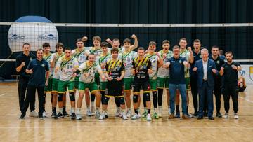 Drużyna siatkarzy w strojach sportowych pozuje do zdjęcia na boisku siatkarskim.