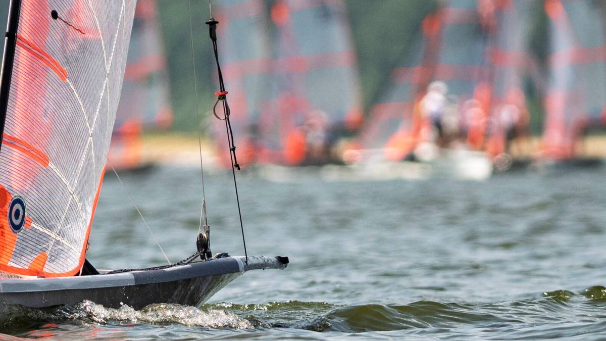 Fragment łodzi żaglowej z pomarańczowo-białym żaglem na pierwszym planie, podczas regat żeglarskich. W tle widoczne rozmyte sylwetki innych łodzi.