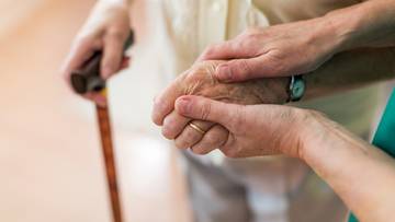 Dłoń wspierająca dłoń osoby starszej, obok znajduje się laska.