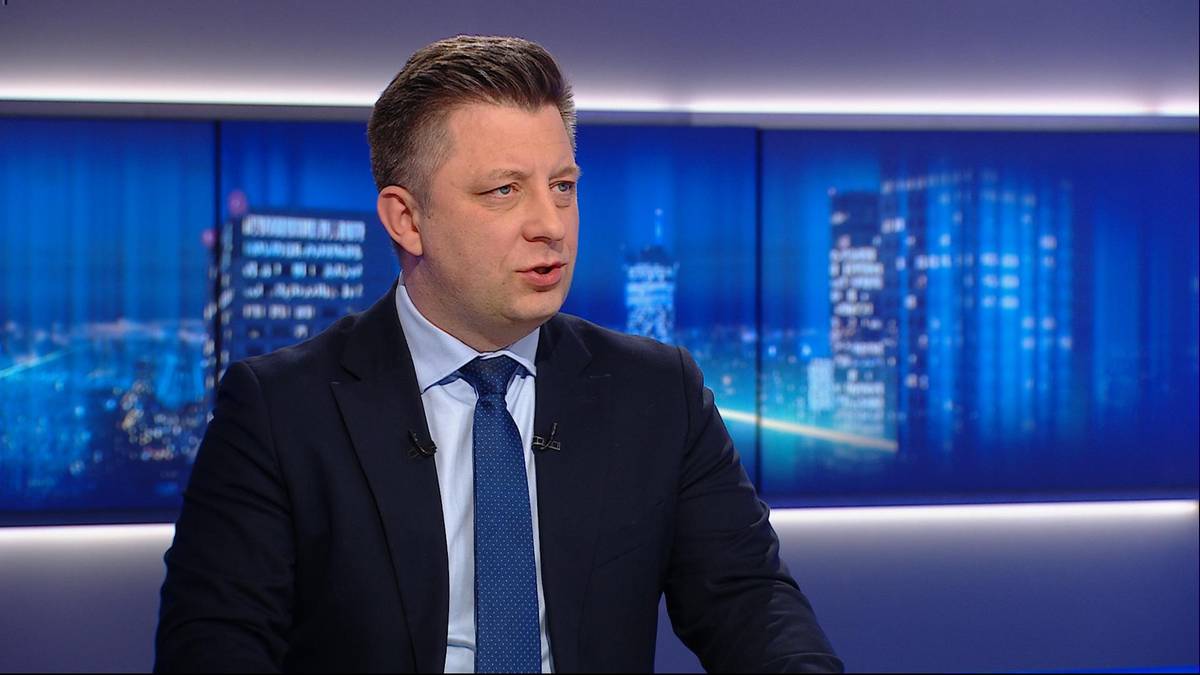 Mężczyzna w garniturze i krawacie m&oacute;wi do kamery, za nim widać ekran z rozmytymi światłami miasta.