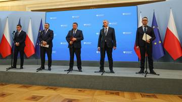 Pięciu mężczyzn w garniturach stoi przed ścianką z polskimi flagami, podczas oficjalnego spotkania lub konferencji.