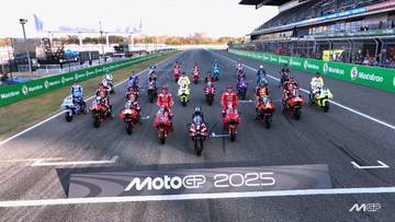 Grupa motocykli MotoGP ustawiona na prostej startowej toru wyścigowego z banerem "MotoGP 2025" na pierwszym planie.