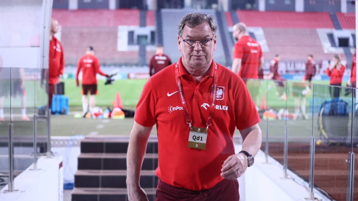 Mężczyzna w czerwonej koszulce z napisem "Polska" i logo Nike, z identyfikatorem na szyi, idzie po schodach na tle stadionu piłkarskiego.