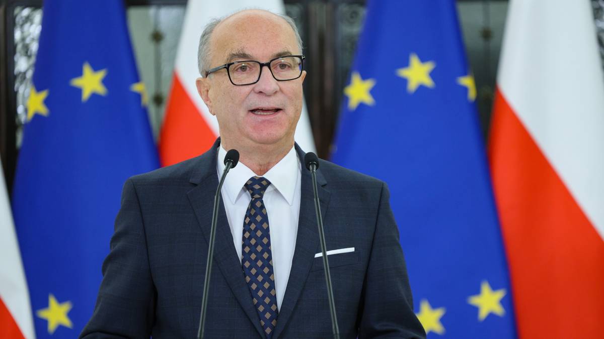 Mężczyzna w garniturze za m&oacute;wnicą, przemawia do mikrofon&oacute;w, w tle flagi Polski i Unii Europejskiej.