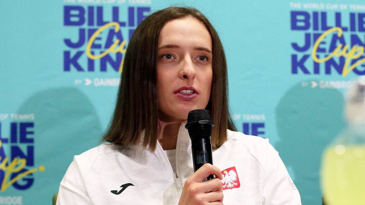 Iga Świątek przemawia do mikrofonu podczas konferencji prasowej, ubrana w białą bluzę z godłem Polski.