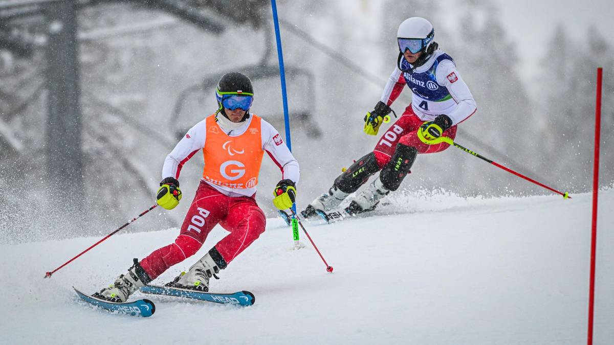Dwóch narciarzy w kombinezonach z biało-czerwonymi flagami Polski, ścigających się na ośnieżonym stoku. Jeden z narciarzy ma na rękawie pomarańczową wstążkę z symbolem paraolimpijskim.