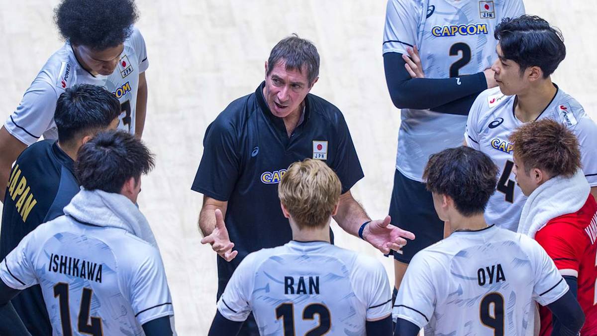 Trener w czarnej koszulce dyskutuje z grupą siatkarzy w białych strojach.