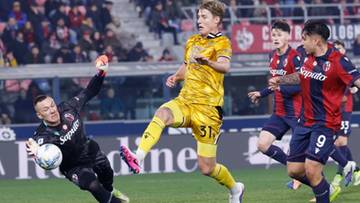 Bramkarz w czarnym stroju rzuca się do obrony, próbując złapać piłkę. Trzech innych zawodników, dwóch w czerwono-niebieskich strojach i jeden w żółtym, znajduje się w pobliżu.