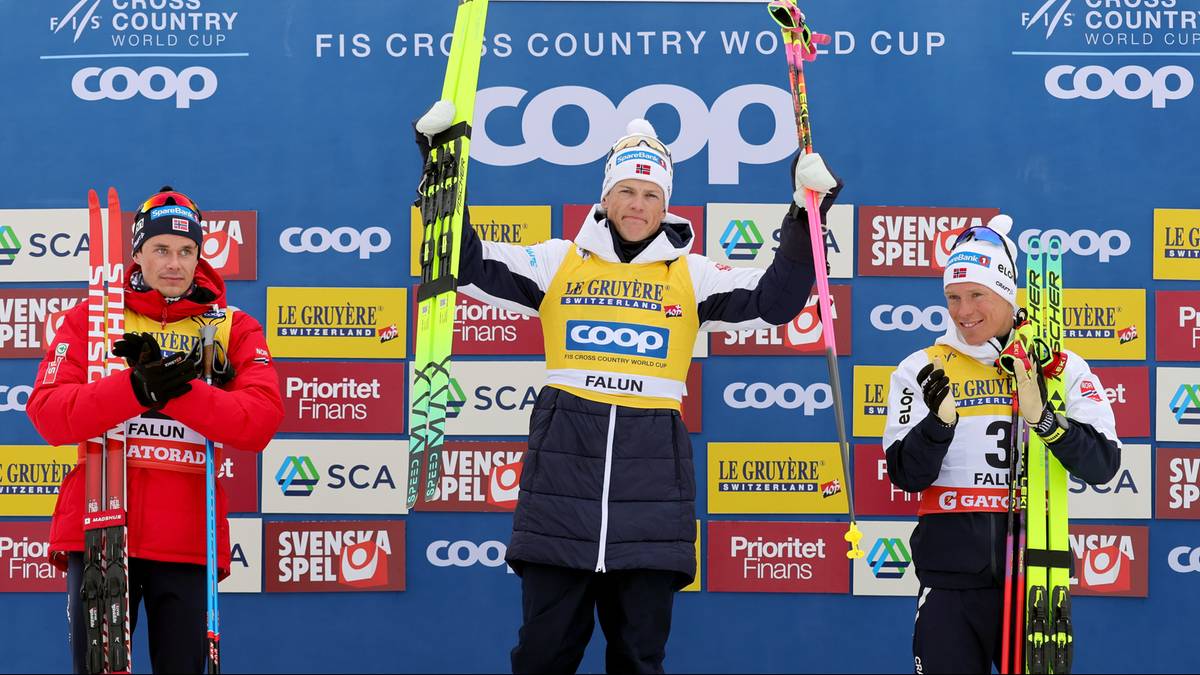 Trzech narciarzy biegowych na podium, z czego środkowy zwycięzca, prezentuje swoje narty i kije.