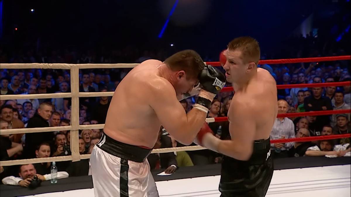 Dwóch bokserów w rękawicach walczy na ringu bokserskim. Jeden z bokserów, w białych spodenkach, klęka, zasłaniając się przed uderzeniem. Drugi bokser, w czarnych spodenkach, przygotowuje się do wyprowadzenia ciosu. Widownia w tle.