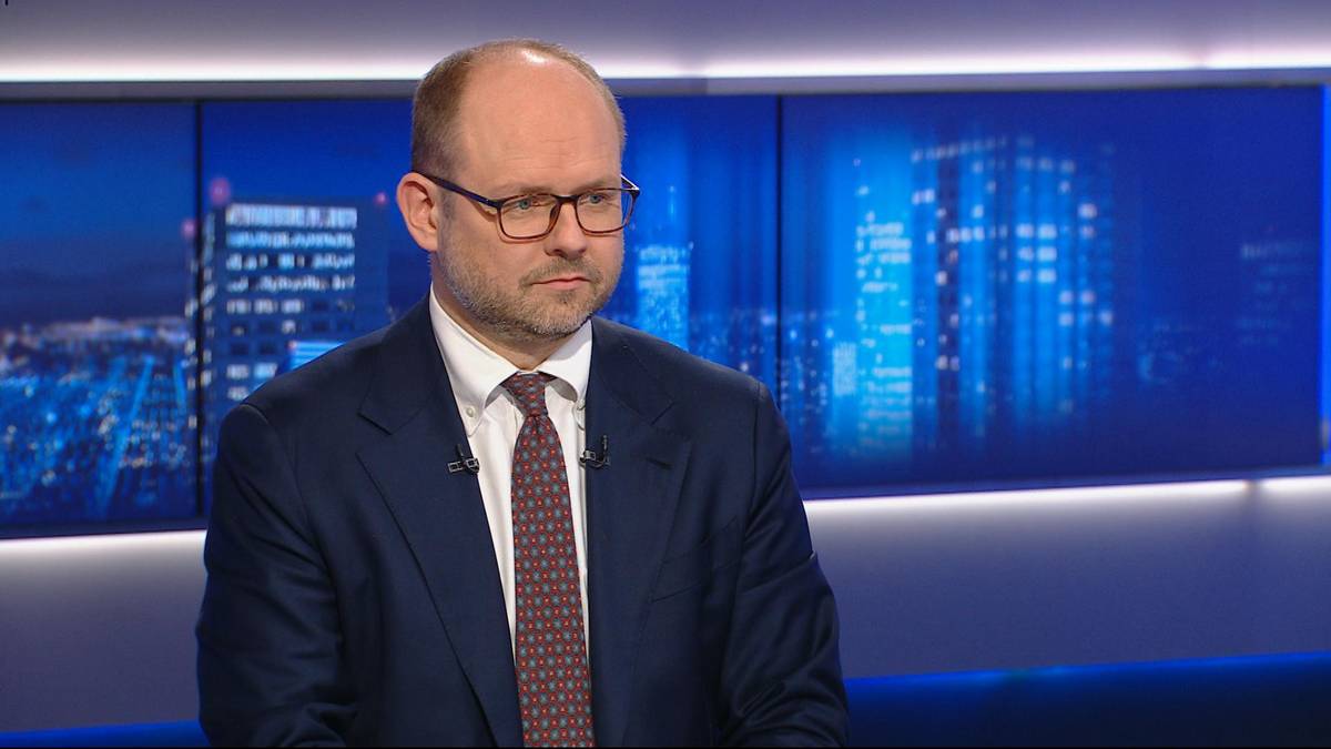 Mężczyzna w garniturze i okularach siedzi przed ekranem z niebieskimi światłami.