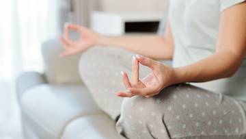 Kobieta w pozycji medytacyjnej na kanapie, z dłońmi złożonymi w mudrę.