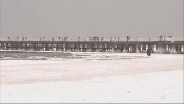 Zamarznięty Bałtyk z widocznym molo w tle, po lodzie spaceruje para.