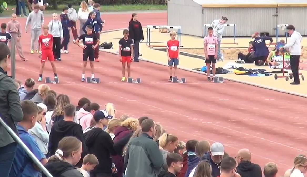 Grupa młodych sportowców stoi w blokach startowych na bieżni stadionu lekkoatletycznego, gotowa do biegu. W tle widać zgromadzonych widzów.