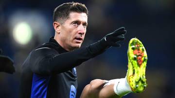 Piłkarz Robert Lewandowski w czarnej koszulce treningowej z niebieskimi rękawami, wykonujący ćwiczenia rozciągające nogę, na której ma żółtą korkową piłkę.