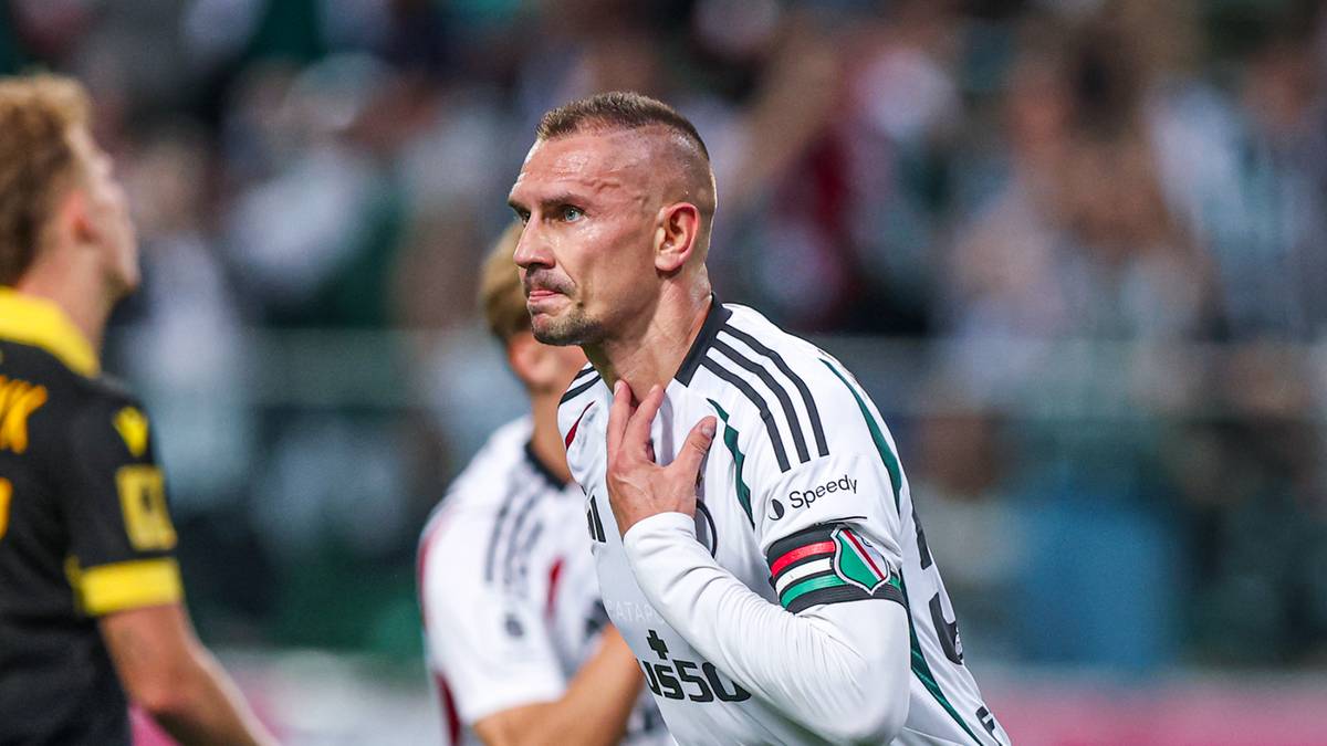 Piłkarz Artur Jędrzejczyk w białej koszulce z czarnymi pasami, gestykuluje prawą ręką przy klatce piersiowej.