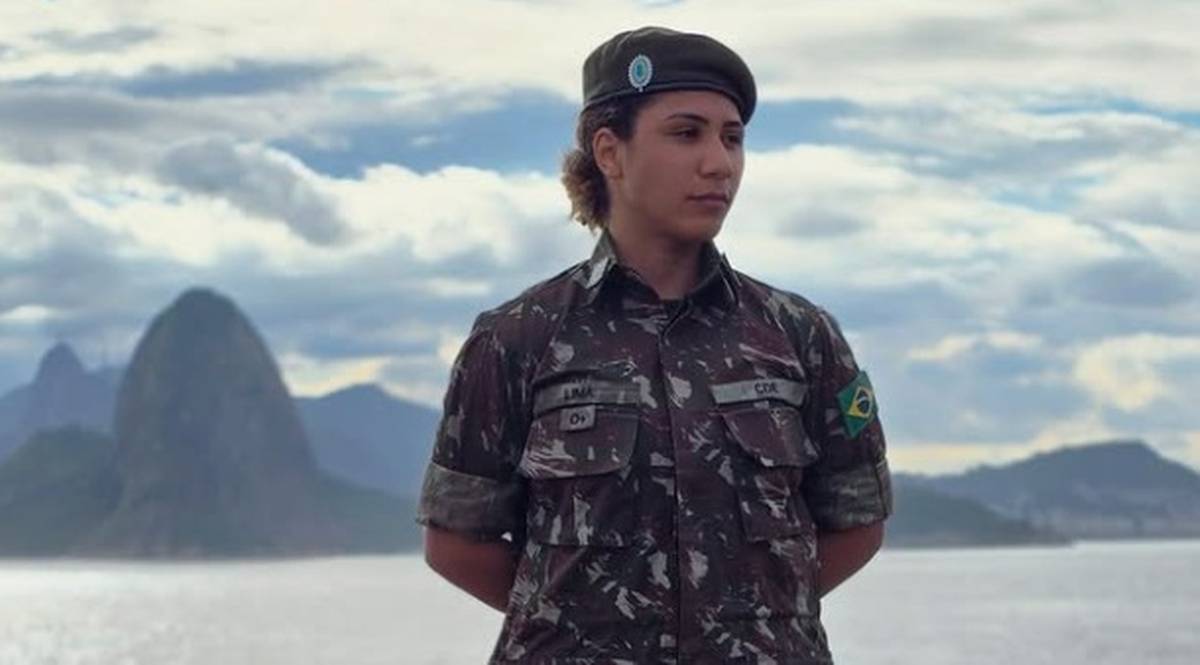 Portret kobiety w mundurze wojskowym i beretce, stojącej przed krajobrazem rzeki i gór.