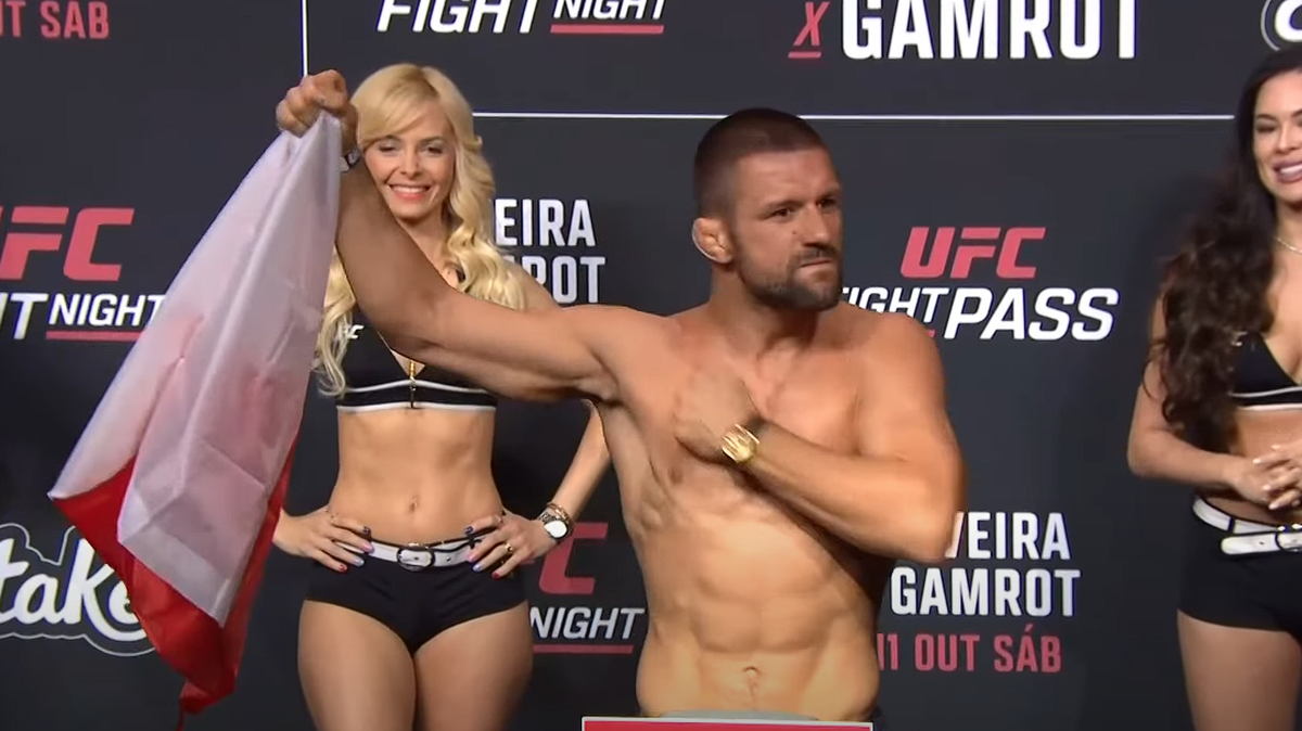 Mateusz Gamrot, zawodnik MMA, stoi na wadze UFC, trzymając polską flagę i patrząc w stronę widowni. Obok niego stoją dwie "ring girls" w strojach sportowych.
