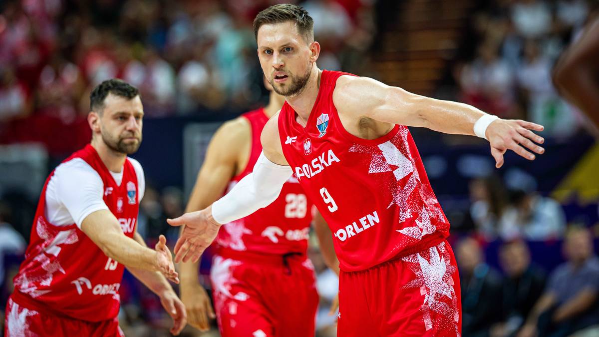 Dwóch polskich koszykarzy w czerwonych strojach, jeden z nich podaje dłoń drugiemu, z widocznymi numerami 9 i 29 na koszulkach. W tle widoczni inni zawodnicy i trybuny.