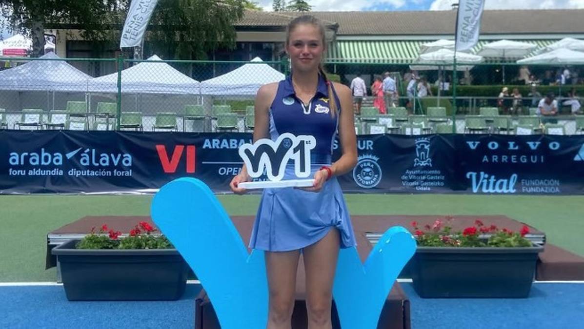 Młoda kobieta, tenisistka, stoi na podium i trzyma nagrodę w kształcie litery 'W'. Jest ubrana w str&oacute;j tenisowy.