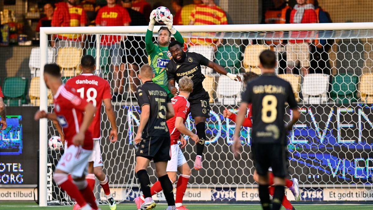 Piłkarze w czarnych i czerwonych strojach walczą o piłkę w polu karnym. Bramkarz w zielonym stroju łapie piłkę, nad głowami ma piłkę, która leci w stronę bramki. Piłkarz w czarnym stroju z numerem 8 na plecach jest po prawej stronie.