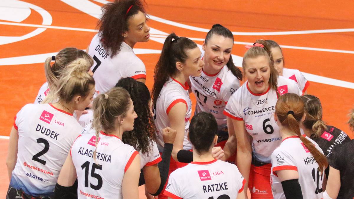 Grupa siatkarek w białych strojach z czerwonymi elementami, trzymających się za ramiona i rozmawiających, podczas meczu Tauron Ligi.