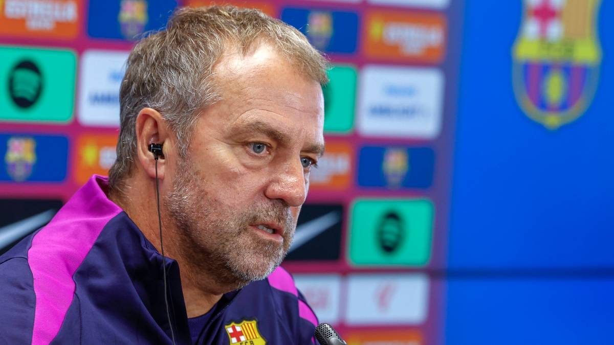 Hansi Flick, trener piłkarski, w ciemnej kurtce z fioletowymi akcentami, z słuchawkami w uszach, mówi podczas konferencji prasowej. W tle widoczny jest niebieski baner z logo FC Barcelony i kolorowymi panelami.