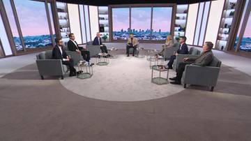 Grupa os&oacute;b siedzi w fotelach w p&oacute;łokręgu w studiu telewizyjnym, dyskutując. W tle znajduje się duże okno z widokiem na panoramę miasta.