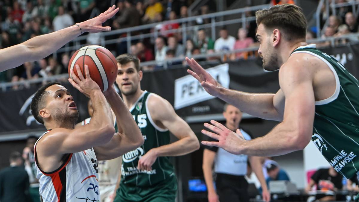 Gracz w białej koszulce z pomarańczową piłką do koszyk&oacute;wki w rękach, gotowy do rzutu, broniony przez gracza w zielonej koszulce.