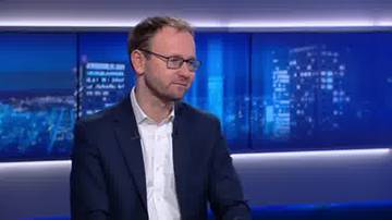 Mężczyzna w okularach i ciemnej marynarce siedzi w studiu telewizyjnym, na tle niebieskich ekran&oacute;w z panoramą miasta.