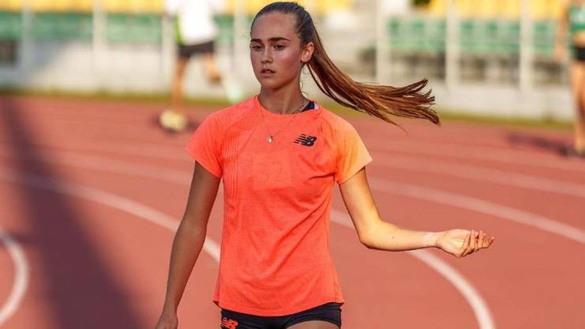 Młoda lekkoatletka w pomarańczowej koszulce z logo New Balance biegnie po bieżni, jej długie włosy są związane w kucyk i falują w ruchu. Obok niej widać fragmenty innych zawodników.