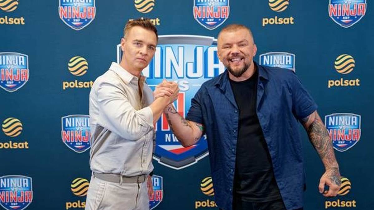 Dw&oacute;ch mężczyzn stoi obok siebie, podając sobie ręce na tle niebieskiej ściany z logo Ninja Warrior Polska.