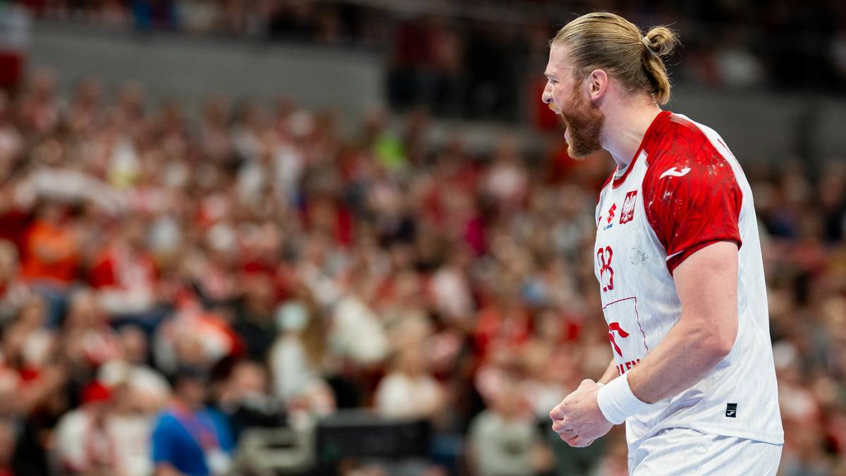 Piłkarz ręczny w białej koszulce z czerwonymi elementami krzyczy podczas meczu, z publicznością na trybunach w tle.