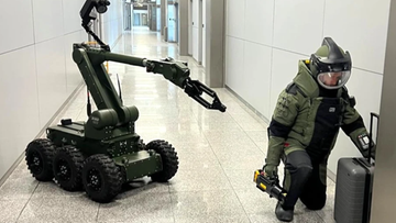 Funkcjonariusz w stroju pirotechnicznym klęczy przy walizce, a obok niego znajduje się robot pirotechniczny w korytarzu lotniska.