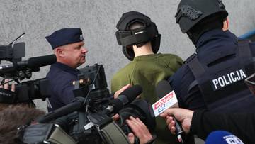 Zatrzymany mężczyzna w hełmie i zielonej bluzie prowadzony przez policjant&oacute;w w otoczeniu dziennikarzy z kamerami i mikrofonami.