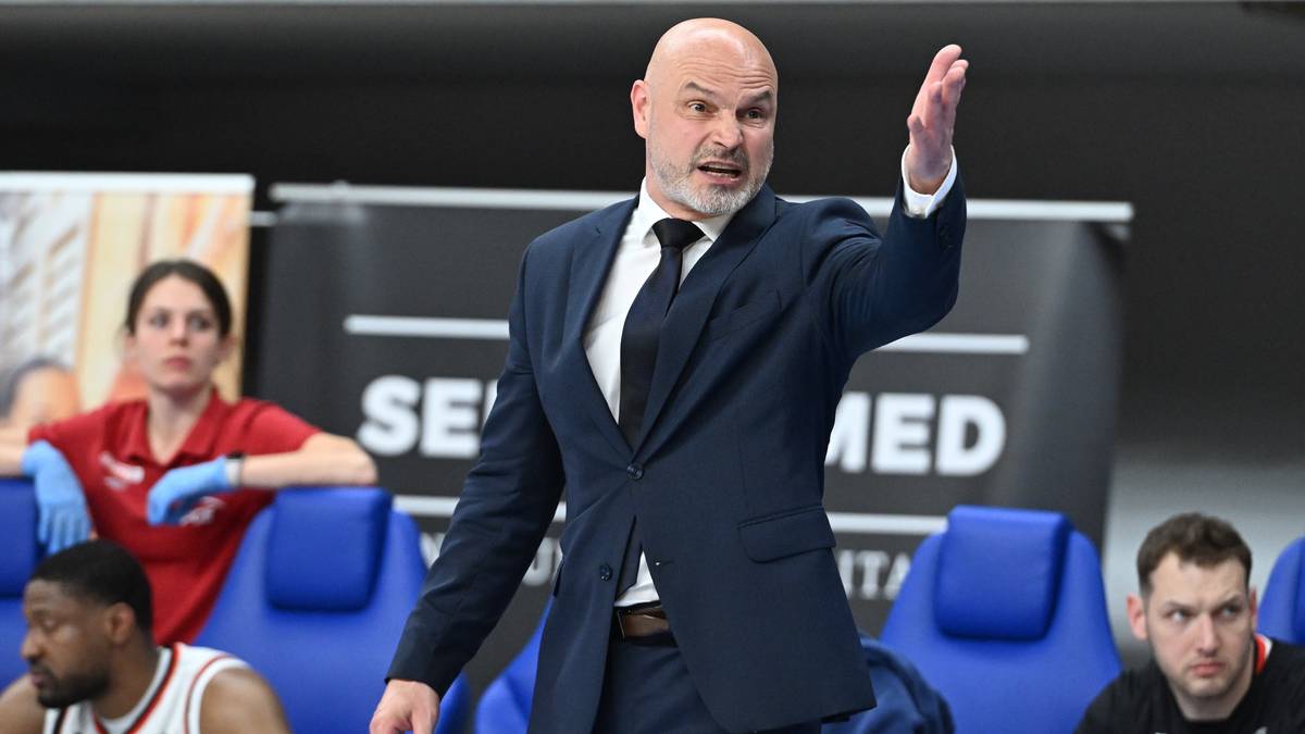 Trener koszykówki w garniturze, z uniesioną prawą ręką, gestykuluje w trakcie meczu.