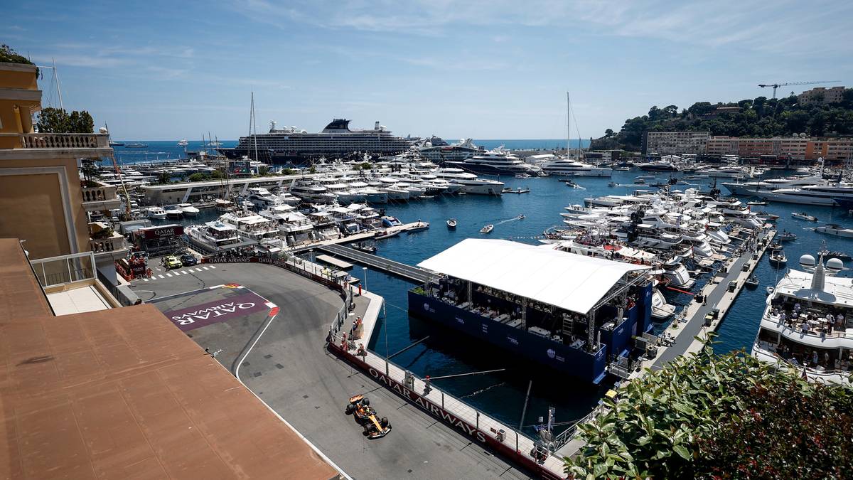 Fragment toru Formuły 1 w Monako, widoczny zakręt z wyznaczonymi liniami i barierami. Po lewej stronie zabudowania, a w tle rozległa marina z licznymi jachtami i statkami.
