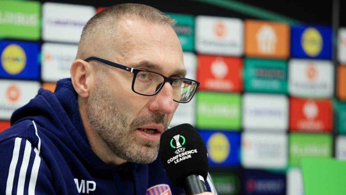 Zbliżenie na twarz mężczyzny w okularach, mówiącego do mikrofonu. Tło stanowi ściana z kolorowych paneli z logo "UEFA Europa Conference League".